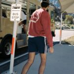 Man wearing red sweatshirt and navy Adidas shorts standing by a bus stop.