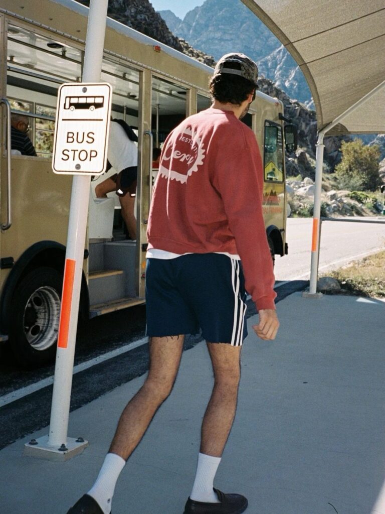Man wearing red sweatshirt and navy Adidas shorts standing by a bus stop.