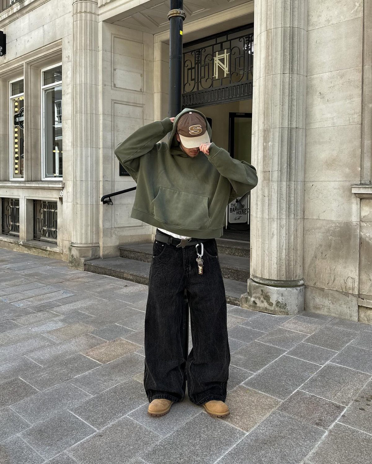 Man wearing an oversized olive green hoodie and wide-leg black trousers on a sidewalk.
