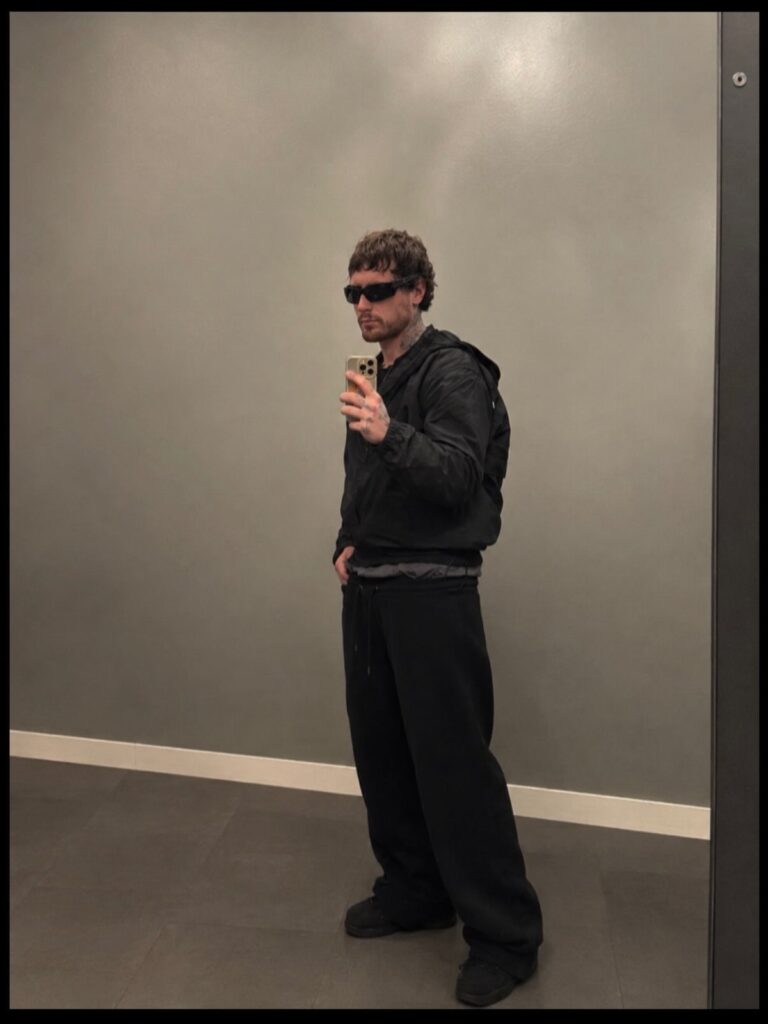 Man in black sunglasses, black windbreaker, and baggy black sweatpants posing for selfie.