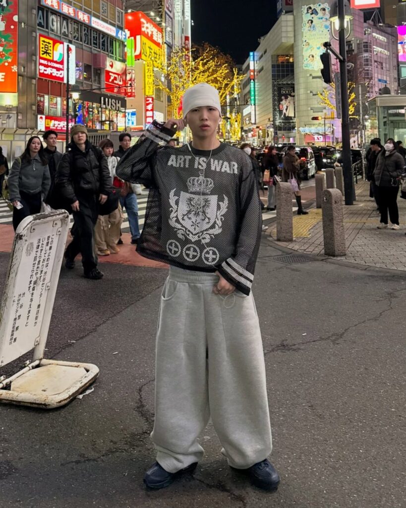 Man in white beanie and oversized grey sweatpants posing on a neon city street.