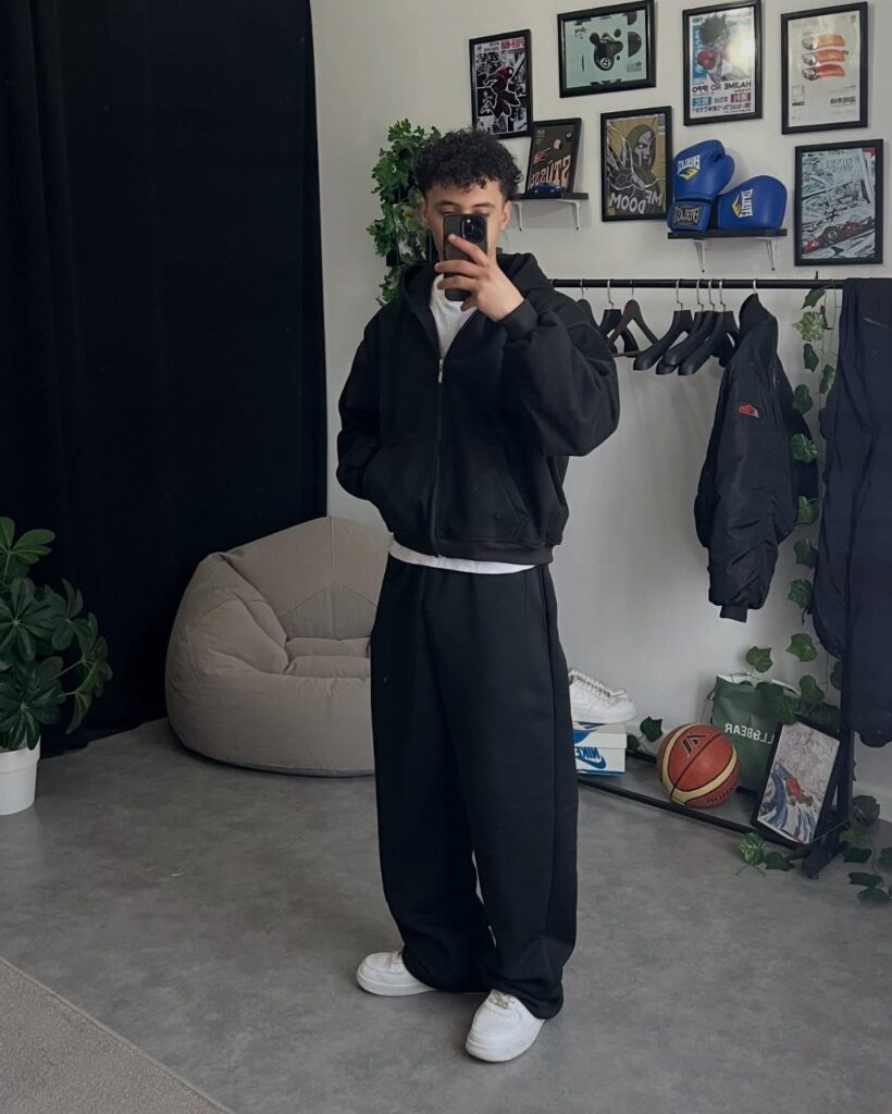 Person wearing baggy black sweatpants, a black jacket, and white sneakers in a room.