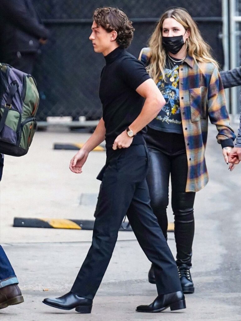Man in all-black outfit and polished black boots walking outdoors with a companion.