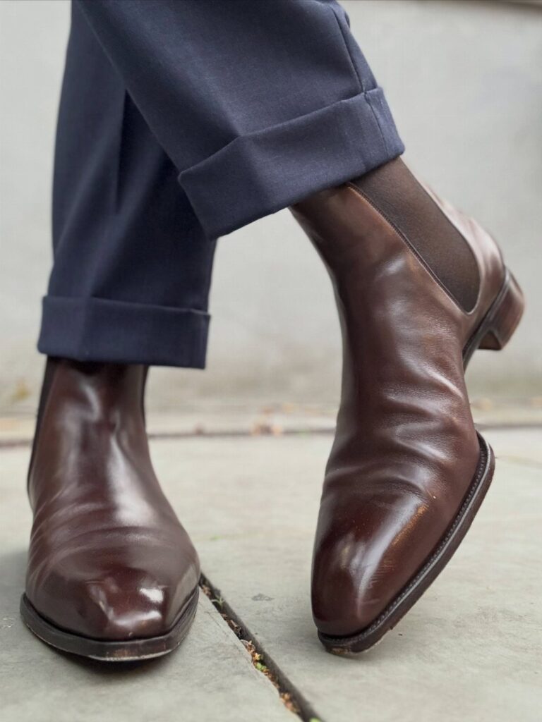 Brown leather Chelsea boots paired with cuffed navy blue trousers on concrete ground.