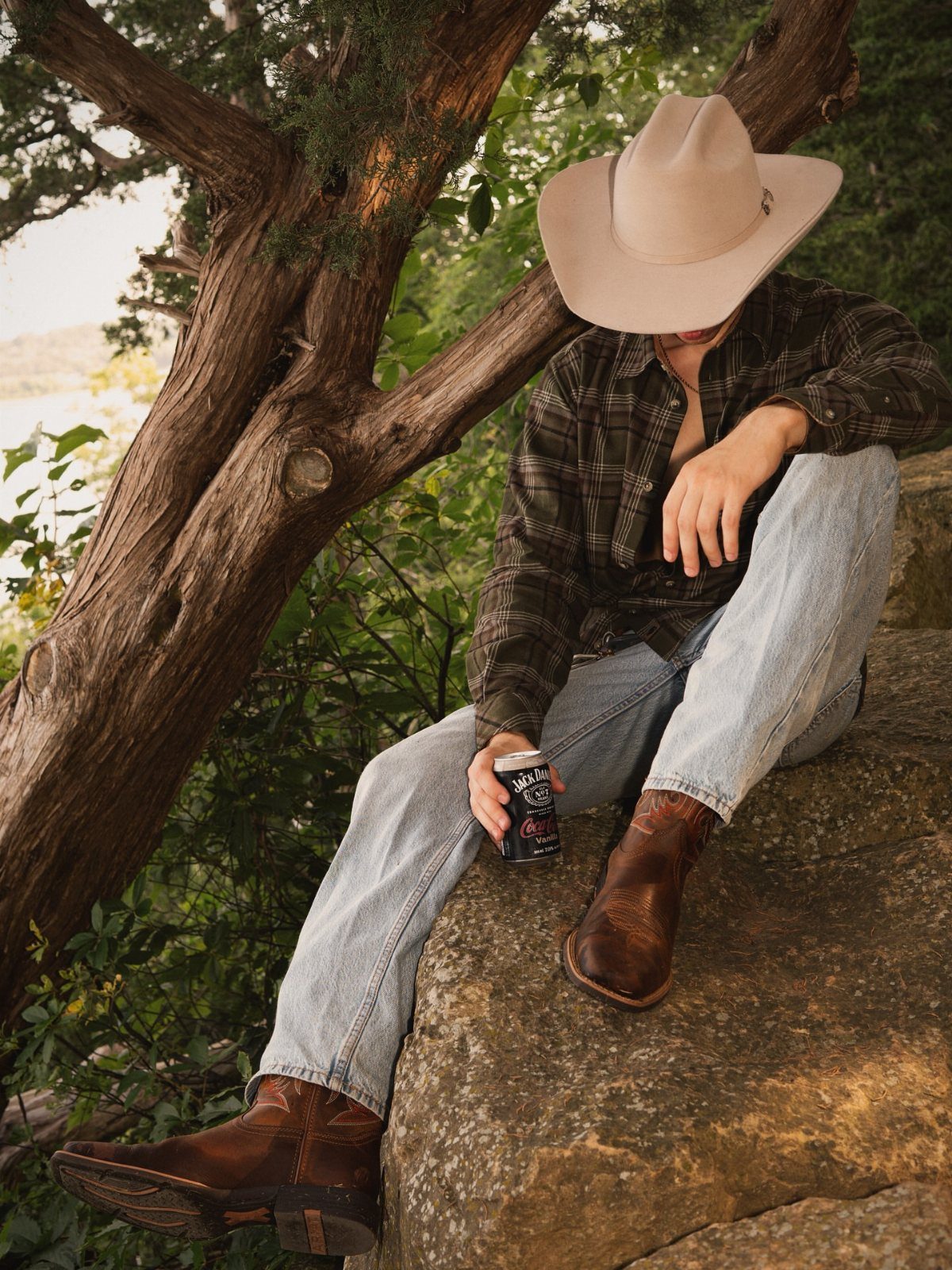 Person in cowboy hat, plaid shirt, light jeans, and brown cowboy boots sitting outdoors.