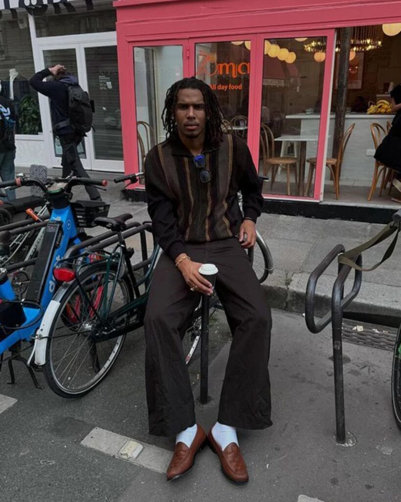 Man wearing brown loafers, dark trousers, and a striped sweater sitting on a street.