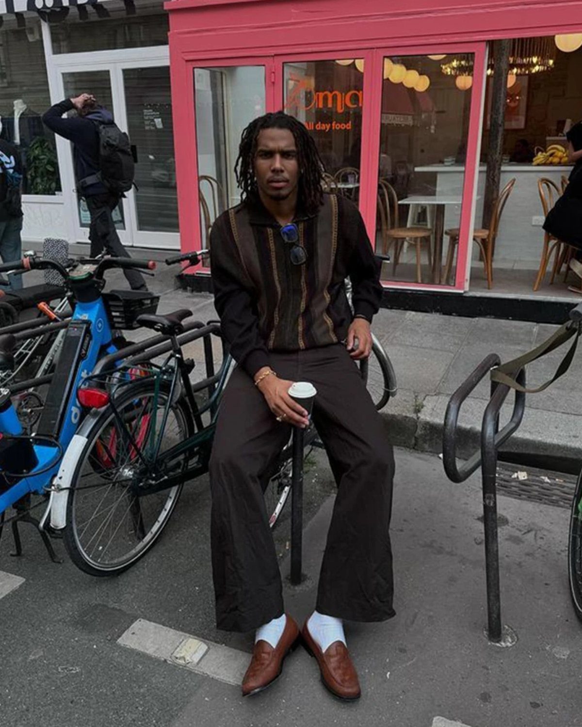 Man wearing brown loafers, dark trousers, and a striped sweater sitting on a street.