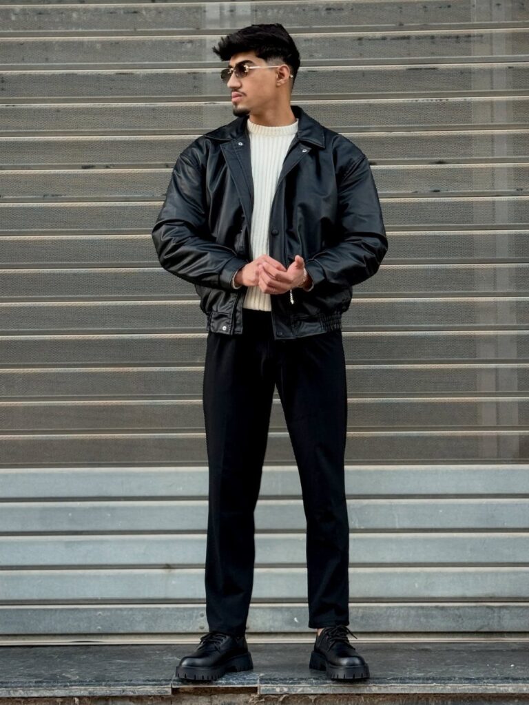 Man wearing black leather jacket, white sweater, black trousers, and chunky black derby shoes.