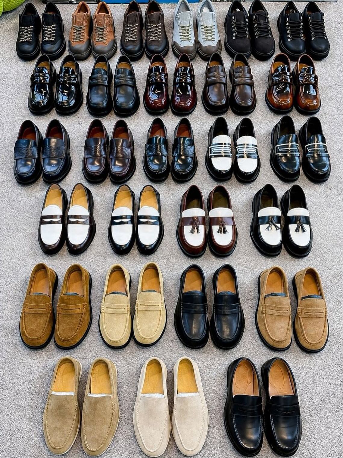 Various men's dress shoes in leather and suede arranged in rows on carpet.
