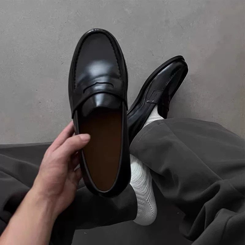 Black leather loafers worn with white socks and dark grey trousers.