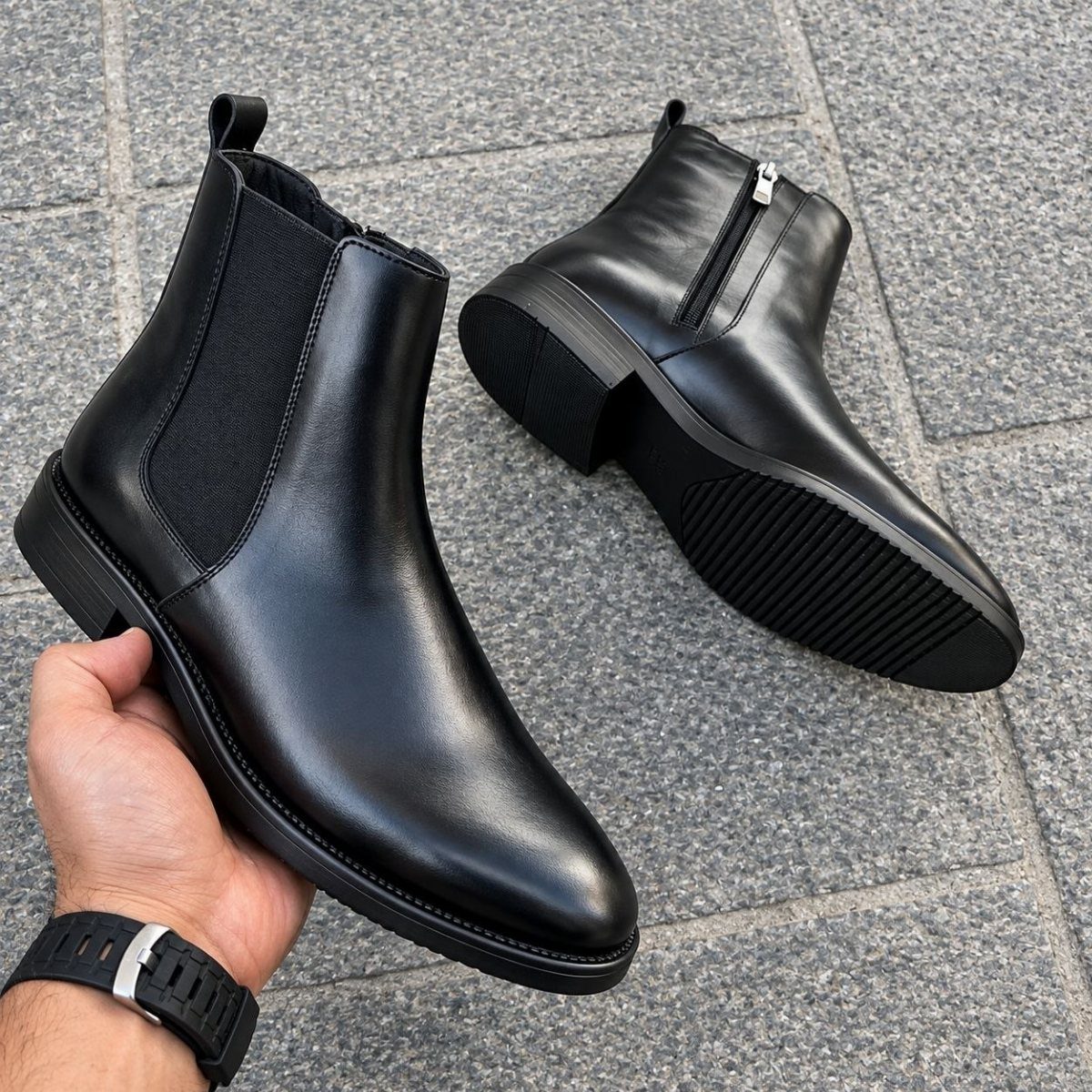 Black leather Chelsea boots with elastic side panels on a grey stone surface.