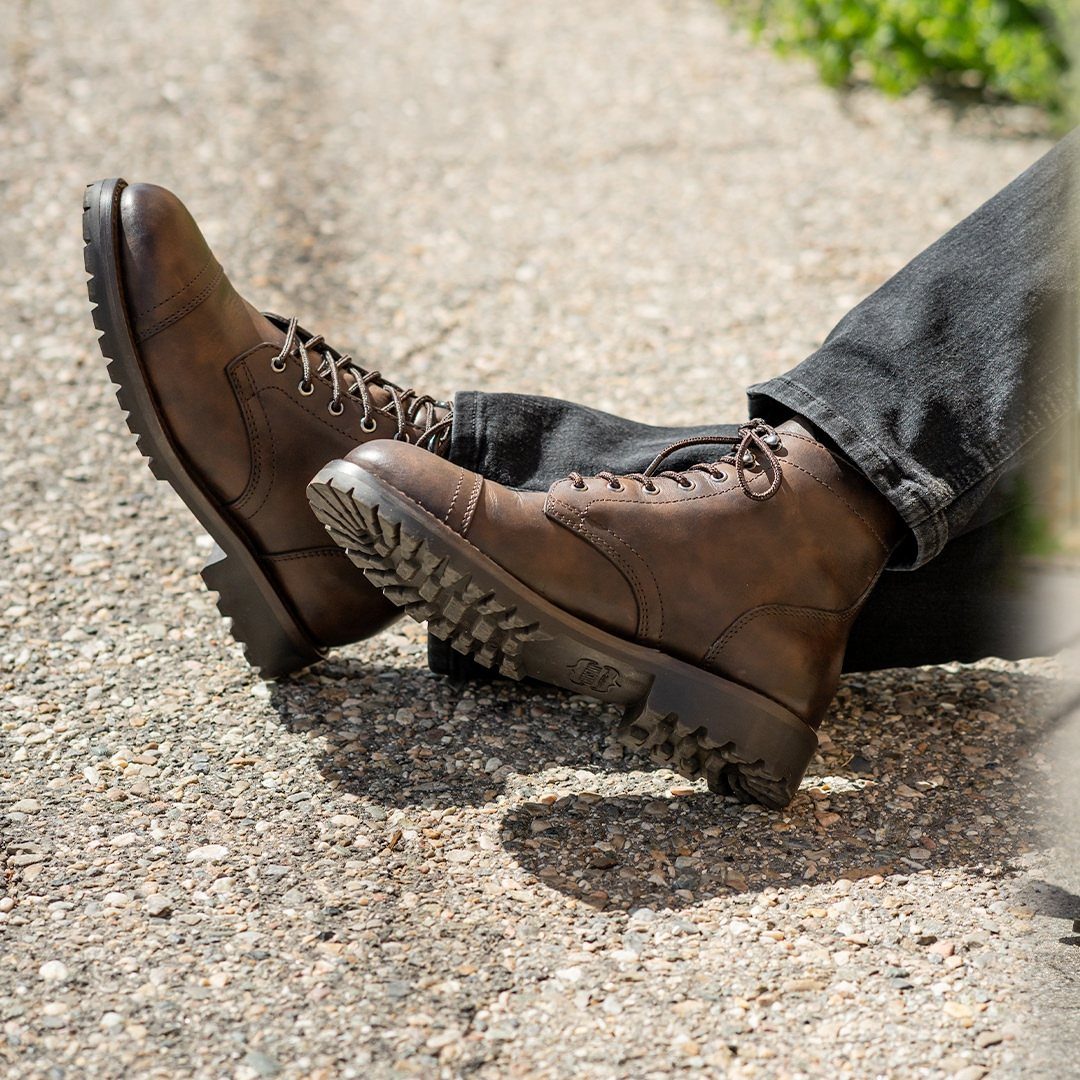 Brown leather lace-up dress boots paired with dark grey straight-leg jeans on gravel.