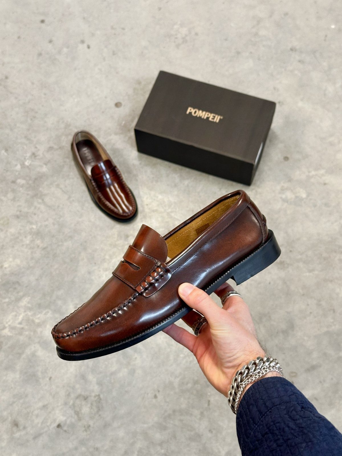 Brown leather penny loafers held by a hand next to a black shoe box.