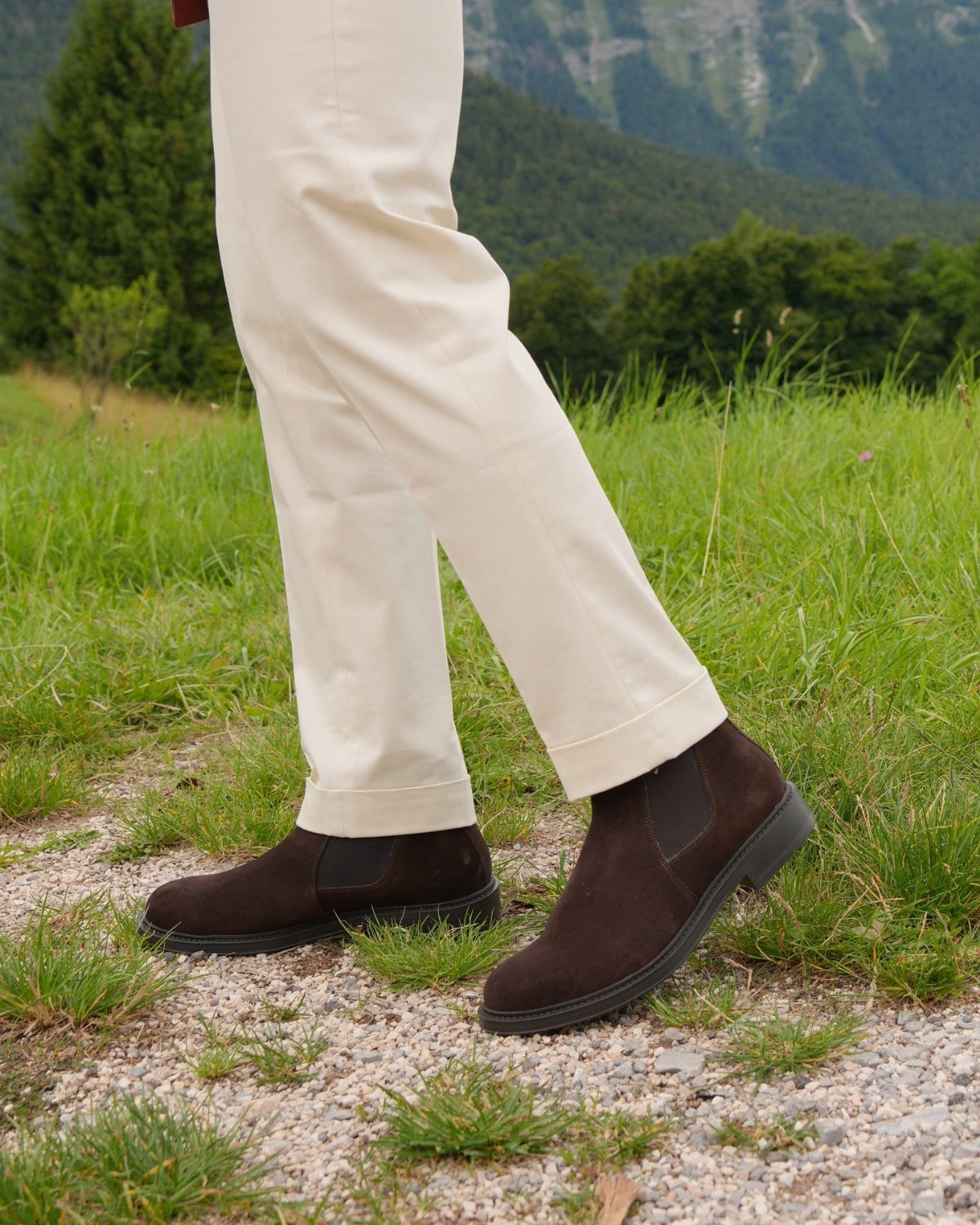 Dark brown suede Chelsea boots paired with cream-colored chinos on a gravel path.
