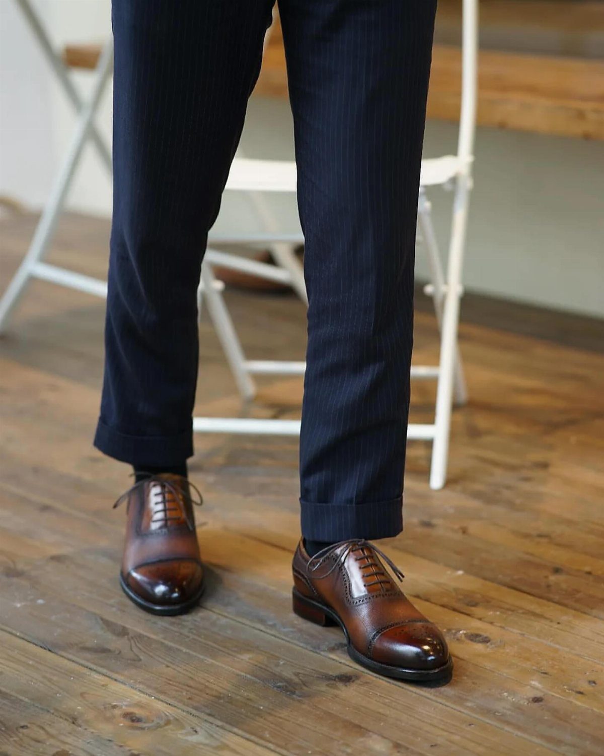 Brown leather cap-toe Oxford shoes paired with navy pinstriped trousers on wooden flooring.