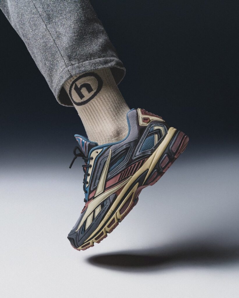 Multi-colored retro running shoe on a foot wearing grey trousers and beige socks.