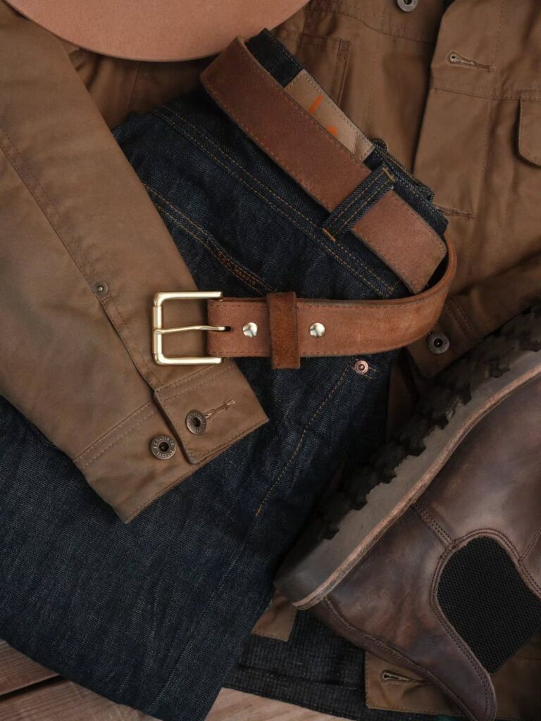 Brown leather belt with gold buckle resting on dark denim jeans and tan jacket.