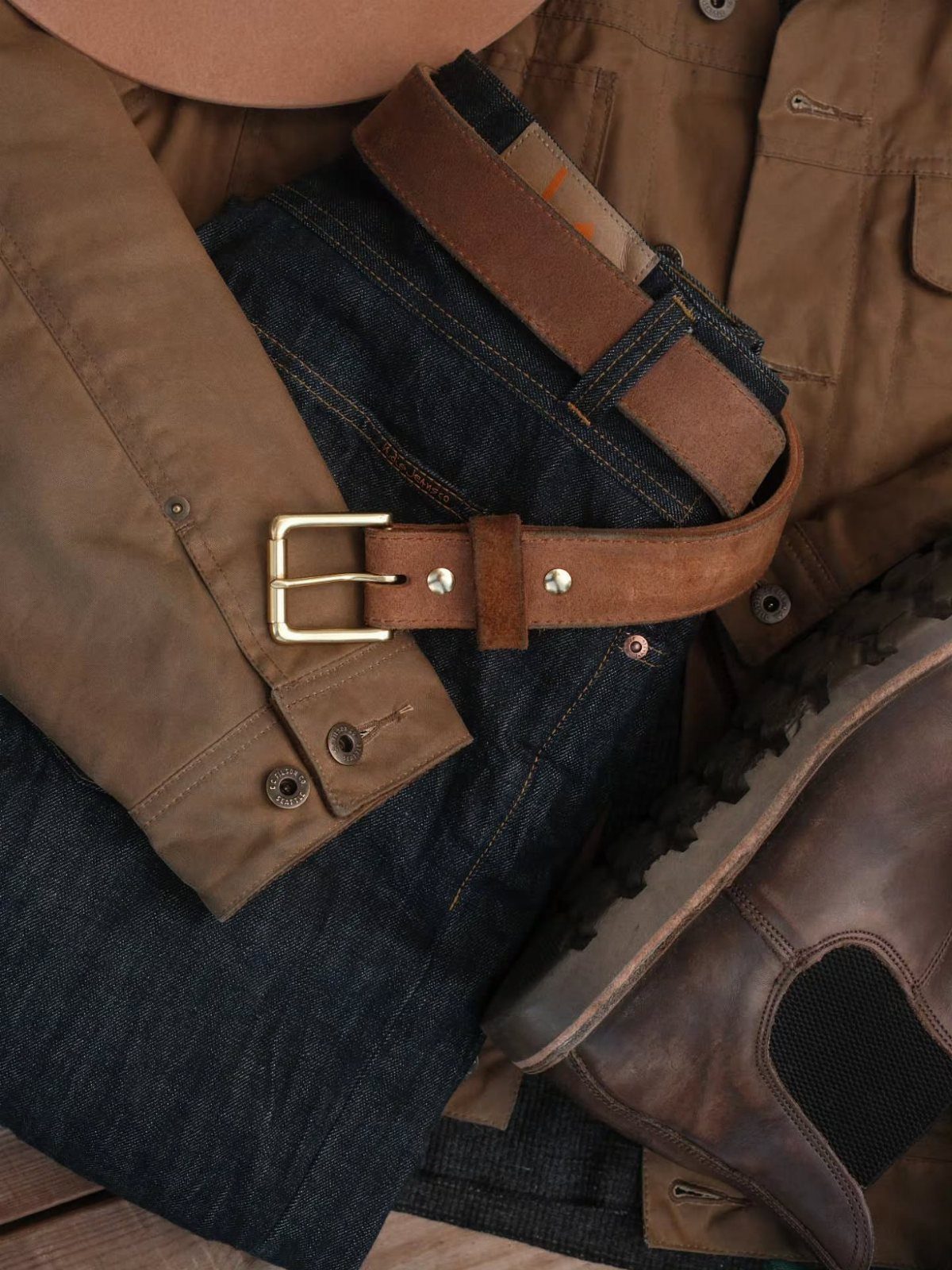 Brown leather belt with gold buckle resting on dark denim jeans and tan jacket.