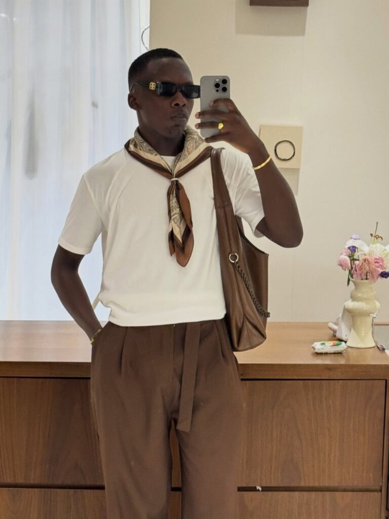 Man wearing a brown patterned scarf, white t-shirt, and brown trousers in mirror selfie