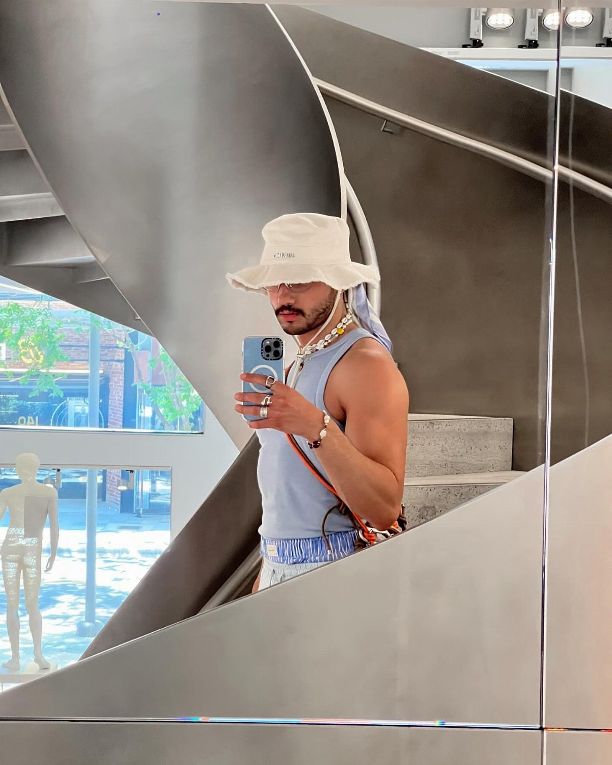 Man wearing a white bucket hat and beaded jewelry taking a mirror selfie.