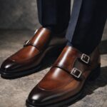 Brown leather double monk strap shoes with silver buckles and navy trousers.