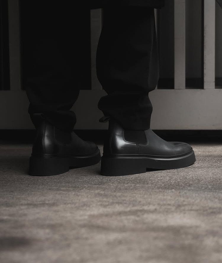 Black chunky leather Chelsea boots worn with black trousers on concrete ground.